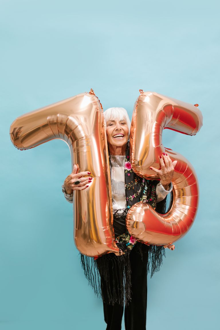 Happy senior woman holding 75 balloons on blue background.
